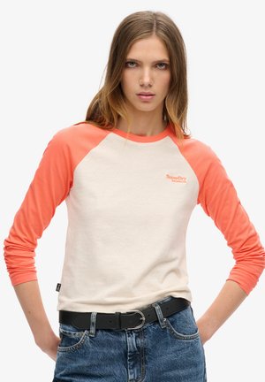 Kvinde med langt lysebrunt hår iført en hvid og koralrød raglan-ærmet skjorte kombineret med blå jeans og et sort bælte, stående med hænderne på hoften.