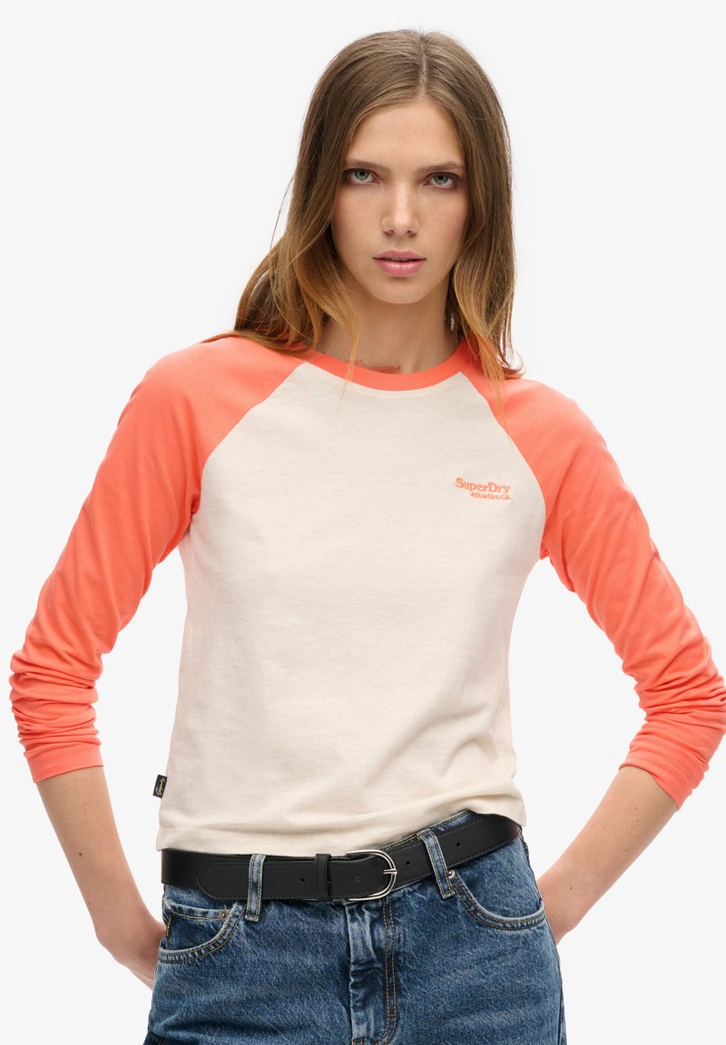 Woman with long light brown hair wearing a white and coral raglan sleeve shirt paired with blue jeans and a black belt, standing with hands on hips.