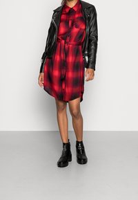 Robe chemise à carreaux rouges et noirs avec des boutons et une ceinture à nouer. Associée à une veste de biker en cuir noir et des bottines noires épaisses.