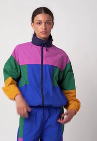 Šareni vjetrovka jakna s ljubičastim, zelenim, plavim i narančastim panelima, patentnim zatvaračem, visokim ovratnikom i elastičnim manžetnama.