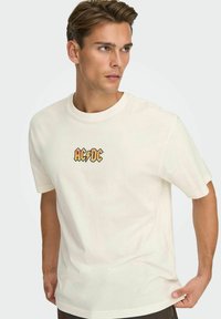 Cream cotton t-shirt with short sleeves, featuring an orange and blue AC/DC logo graphic on the chest. Classic crew neckline design.