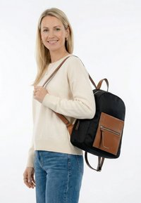 Smiling woman wearing cream sweater and blue jeans carries black backpack with brown leather front pocket and straps on her shoulder.