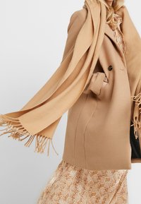 Manteau en laine beige à coupe ajustée, avec deux boutons et une poche. Complété par une écharpe assortie à franges, porté sur une robe à motifs.