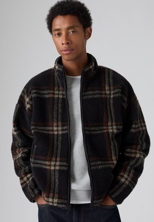 Hombre con una chaqueta de forro polar a cuadros en negro y marrón sobre una camiseta gris claro con las manos en los bolsillos frente a un fondo liso.