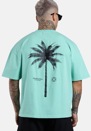 Mann mit Hals-Tattoo und Sonnenbrille trägt ein mintgrünes T-Shirt mit großem schwarzem Palmenmotiv auf dem Rücken.