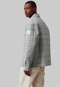 Graue gesteppte Jacke mit Kragen und Knopfverschlüssen, ausgestattet mit einem Logo-Patch am linken Arm und Kontrastnähten.