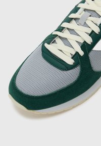 Der Sportschuh verfügt über ein Obermaterial aus grünem Wildleder, grauem Mesh und weißen Schnürsenkeln. Die Sohle ist leicht und hat eine leicht strukturierte Oberfläche.