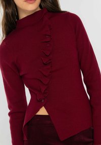 Suéter burdeos con cuello alto y tapeta abotonada con volantes, con mangas largas y dobladillo asimétrico.