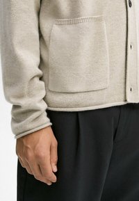 Beige gebreide cardigan met een gestructureerd oppervlak, knoopsluiting en een opgestikte zak aan de linkerkant, gecombineerd met zwarte broeken.