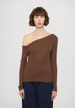 Brown ribbed long-sleeve top with one shoulder exposed; soft texture, form-fitting design, and straight hem. Worn with dark blue trousers.