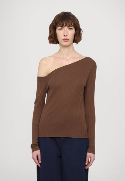 Haut long à manches ribbées marron avec une épaule dénudée ; texture douce, design ajusté et ourlet droit. Porté avec un pantalon bleu foncé.