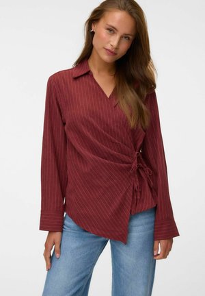 Femme aux longs cheveux bruns portant un chemisier cache-cœur bordeaux à fines rayures et un jean bleu, debout devant un fond uni.