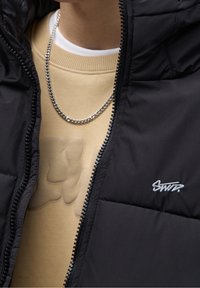 Giacca a vento nera con una superficie lucida e texture. Presenta una chiusura con zip e un piccolo logo ricamato sul petto, indossata sopra un sudore beige.