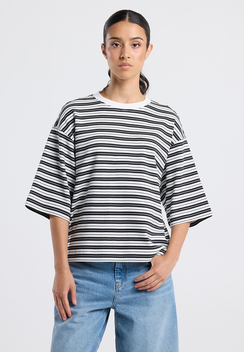 Vrouw in een oversized zwart-witte gestreepte shirt met horizontale strepen en losse lichtblauwe spijkerbroeken, staand tegen een effen achtergrond.