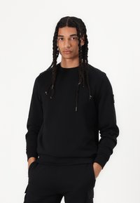 Sort sweatshirt med rund hals, lange ærmer og ribbede manchetter. Har en sidelomme med hardware-detaljer og en glat tekstur.