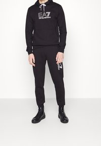 Svart huvtröja med EA7-logotyp på bröstet, i kombination med svarta joggers med logotyp på sidan. Modellen bär svarta snörskor. Slät textur.