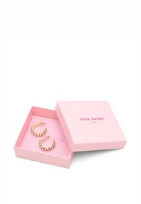 Guldøreringe med hvidt gemstone-accents, vist i en lyserød æske med "TONE DAMILI by A&C" trykt indeni.