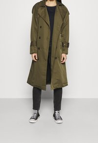 Oliwkowy płaszcz trench z podwójnym zapięciem, dużymi klapami i guzikami, noszony na ciemnych dżinsach i czarnych sneakersach.