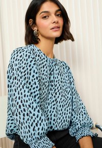 Blå blus med svart prickig mönster, samlade ärmar med elastiska manschetter, lätt material och rund halsringning, stylad med svarta byxor.