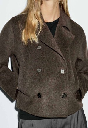 Kobieta w ciemnobrązowym, dwurzędowym, wełnianym płaszczu z guzikami, ręce w kieszeniach, czarna bluzka pod spodem, na jednolitym tle.