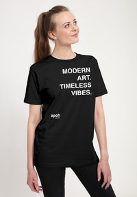 Žena s dlouhým copem, oblečená v černém tričku s bílým textem "MODERNÍ UMĚNÍ. NADČASOVÉ VIBRACE." a malým logem "apoh London", dívající se doprava.