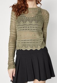 Olivengrüner, gestrickter Crop-Top mit aufwendigen Durchbrüchen; gerippter Ausschnitt und gewellter Saum. Kombiniert mit einem schwarzen, gefältelten Rock.