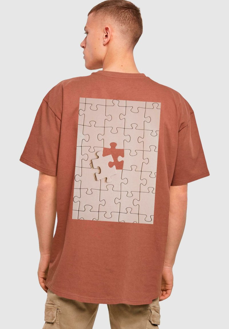 Brun T-shirt med et grafikmotiv af et puslespil på ryggen, hvor en manglende brik er fremhævet i en lys farve og omkranset af sort.