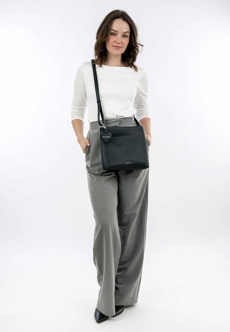 Black leather crossbody bag with a structured shape, featuring a heart-shaped charm and a smooth texture. Styled with gray wide-leg pants.