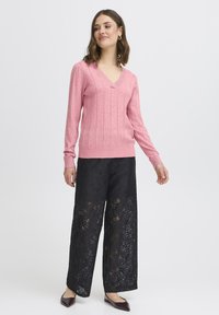 Vállig érő barna hajú nő, rózsaszín V-nyakú pulóvert, fekete csipkés, bő szárú nadrágot és sötét, hegyes orrú cipőt visel, fehér háttér előtt áll.