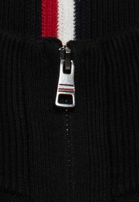 Pull zip noir avec une texture côtelée, fermeture éclair argentée et une bande tricolore rouge, blanche et bleue sur le devant.