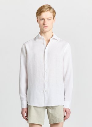 Giovane uomo con capelli chiari che indossa una camicia bianca a maniche lunghe abbottonata e pantaloncini beige a righe, in piedi contro uno sfondo chiaro e uniforme.