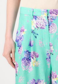 Bukser med blomstermønster i lys teal-stoff som har klynger av lilla og hvite hortensiaer. Inkluderer frontplisseringer og sidelommer.