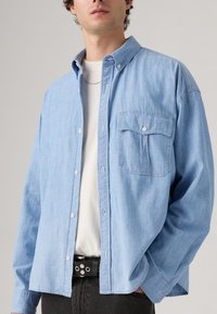 Lyseblå, skjorte med knapper og brystlomme, lavet af et blødt, vævet materiale; båret over en hvid T-shirt og parret med sorte bukser.