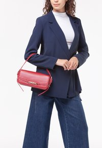 Borsa a tracolla rossa in pelle di forma rettangolare strutturata, con accenti in hardware dorato e tracolla removibile. Indossata con un blazer blu navy e jeans.
