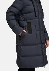 Cappotto imbottito navy con vita aderente, tasche laterali con zip e colletto alto. Presenta un tessuto testurizzato e cinture nere regolabili.
