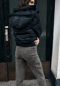 Czarna pikowana kurtka puchowa z wysokim kołnierzem, pikowanym wzorem i zamkami błyskawicznymi; w zestawie z szarymi spodniami cargo z kieszeniami i teksturowanym wykończeniem.