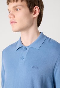 Šviesiai mėlynas polo marškinėliai, pagaminti iš tekstūruotos medžiagos. Turi apykaklę, du mygtukus ir išsiuvinėtą "BOSS" logotipą ant krūtinės.