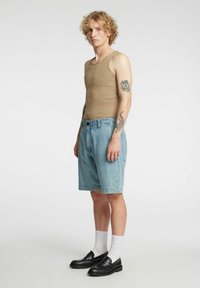 Beige ermeløs topp, lyseblå denimshorts, svarte loafers og hvite sokker. Har krøllete hår og synlige tatoveringer på armen.