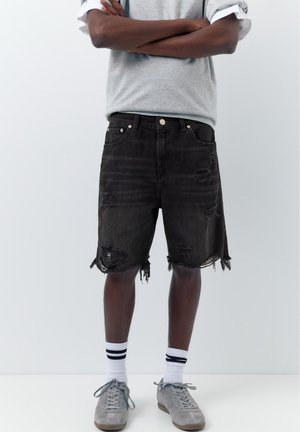 Osoba nosząca czarne, zniszczone szorty jeansowe, szare trampki, białe skarpetki z czarnymi paskami oraz szary krótki rękaw z wywiniętymi rękawami.