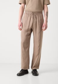 Pantalons relaxes couleur brun clair avec une taille élastique et un cordon de serrage ; tissu lisse et doux avec des coutures latérales, associés à des chaussures noires.
