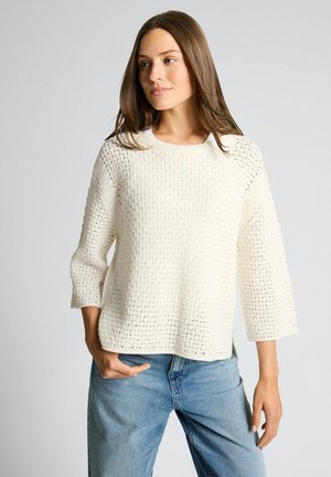 Vrouw met lang bruin haar, gekleed in een gestructureerde gebroken witte gebreide trui en lichtblauwe jeans, poserend met één hand in de zak.