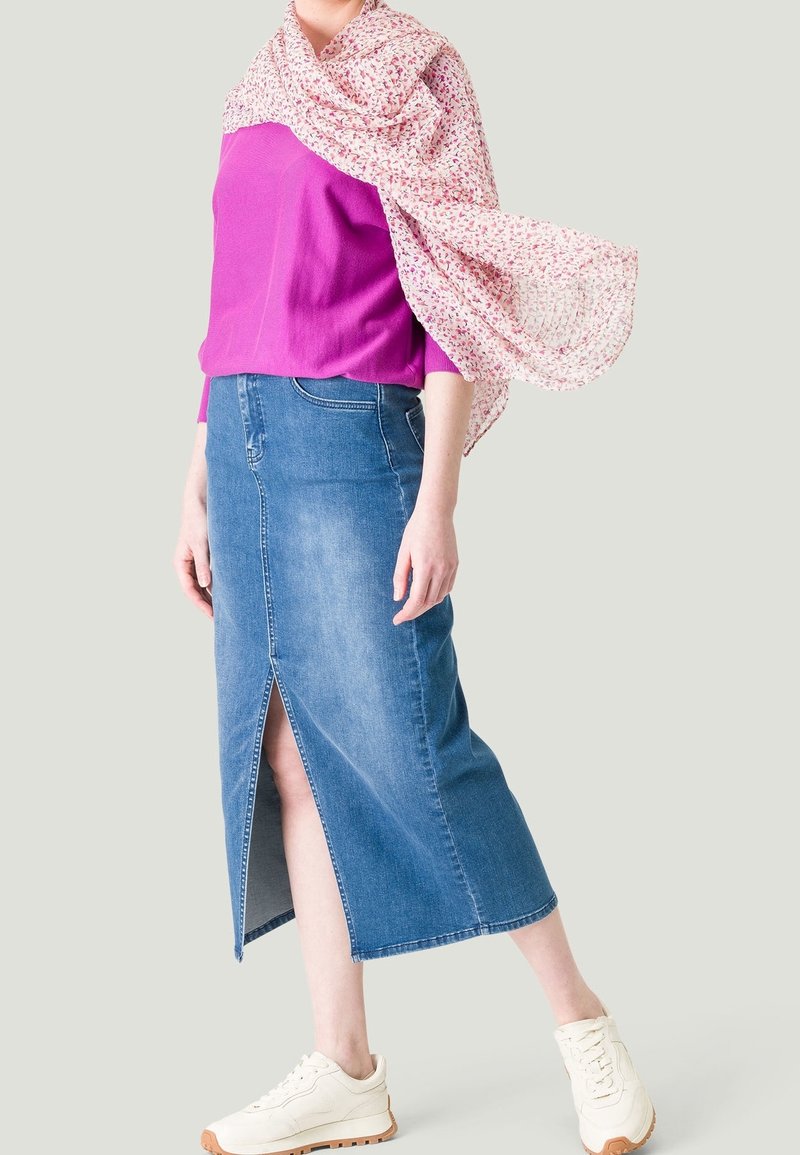 Femme portant un haut rose, une écharpe fleurie flottant dans le vent, une jupe midi en denim avec une fente à l'avant et des baskets blanches sur un fond uni.