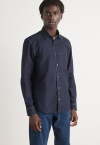 Dark blue button-up shirt with a single chest pocket, contrasting white stitching, and a collared neckline, paired with blue jeans.