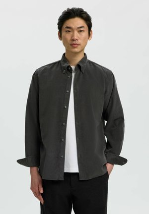 Junger Mann steht frontal, trägt ein dunkelgraues Hemd mit Knöpfen über einem weißen T-Shirt und schwarze Hose, vor einem schlichten hellen Hintergrund.
