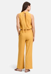 Mosterdgele jumpsuit met een halternek ontwerp, een strikdetail op de rug, wijd uitlopende broekspijpen en een zachte, textuurstof. Slanke pasvorm bij de taille.