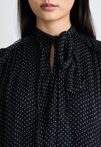 Schwarze Bluse mit einem weißen Punkte-Muster, einer Schleifenkragen und gefältelter Stoffverarbeitung am Ausschnitt und den Schultern.