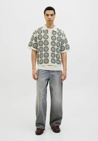 Majica s kratkimi rokavi ima mrežni vzorec črnih in belih cvetlic na kremni podlagi; kombinirana z svetlim širokima kavbojkami in rjavimi čevlji.