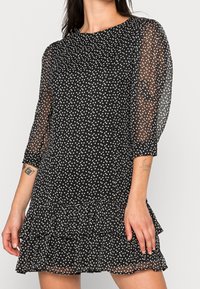 Schwarzes Kleid mit weißen Punkten, transparenten, aufgepufften Ärmeln, rundem Ausschnitt und einem Rüschensaum. Hergestellt aus leichtem, strukturiertem Stoff.