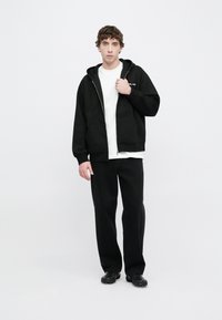 Sudadera negra con cremallera y bolsillos delanteros, llevada sobre una camiseta blanca. Combinada con pantalones negros de corte holgado y calzado negro.