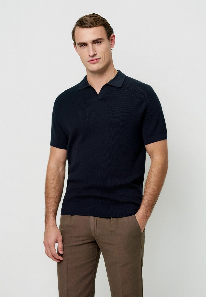 Marineblaues kurzärmliges Poloshirt mit Rippstruktur, umgeschlagenem Kragen und geradem Saum, kombiniert mit hellbraunen Hosen.